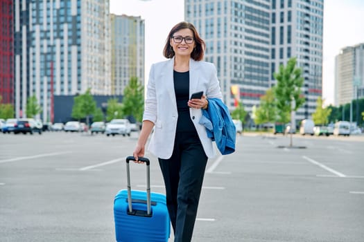 Mature confident business woman walking with suitcase, city background. Smiling middle agged woman with smartphone luggage, looking at camera. Business trip, travel, city, work, lifestyle, people 40s