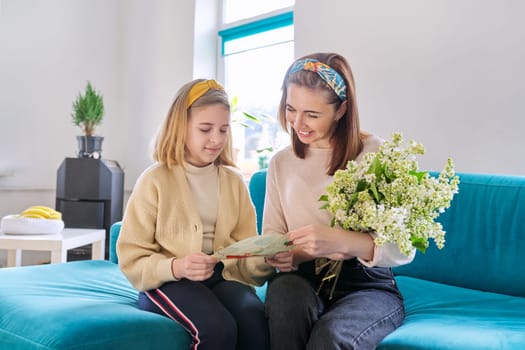 Happy mother and daughter child congratulating with bouquet of flowers and hand-drawn card, at home in living room on sofa. Mother's day, family, relationship, emotions, love, togetherness concept