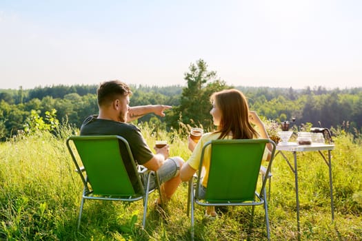 Happy middle aged couple relaxing together outdoor sitting on folding chairs at camping table. Man and woman talking, drinking fresh coffee, enjoying summer nature of wild meadow, back view, weekend