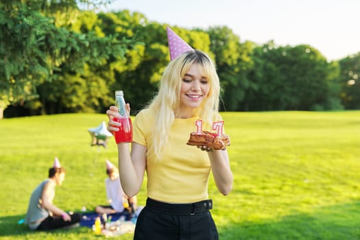 Beautiful teenage girl in festiv hat on birthday with cake and candles. Happy blonde female at outdoor picnic party holding cake with candles 17. Adolescence youth age beauty, holiday birthday concept