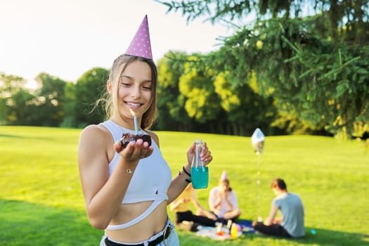 Birthday, teenage girl in festiv hat with cake and candle at outdoor party. Picnic in nature, happy having fun female posing looking at camera. Adolescence, holiday, birthday, celebration, age