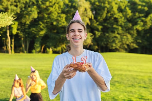 Happy guy teenager in birthday hat with cake with candles 17. Outdoor picnic party with friends in park on grass. Adolescence, having fun, holiday, celebration, birthday, age concept