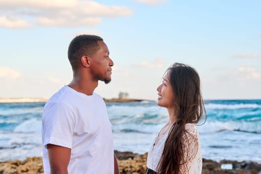 Young beautiful couple looking in the eyes in profile, on seascape. Sky sea background, multicultural couple, Asian woman and African American man. People, relationships, family, happiness, tourism