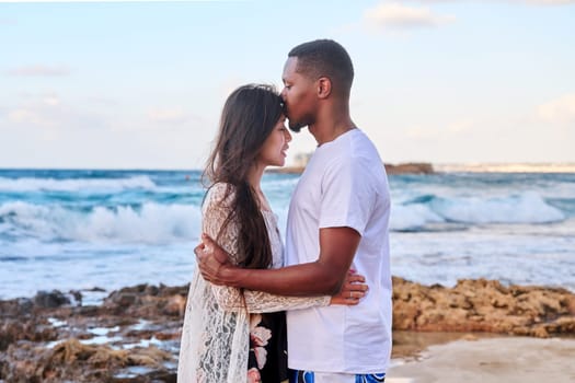 Kiss of loving young couple, sea sky background. Beautiful multicultural couple, profile view, honeymoon at seaside resort, vacation together. Love, relationship, tourism travel, nature beauty people