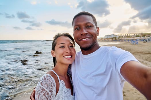 Happy couple in love taking selfie together on smartphone on beach, looking at camera. Vacation tourist trip to seaside resort, summer vacation, multicultural family, happiness, date, love concept