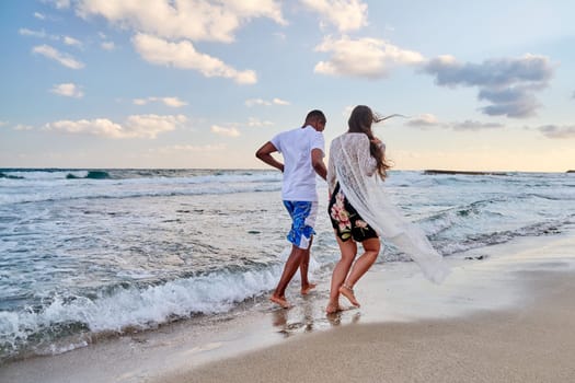 Happy couple running together on the water, on the beach on a summer day, back view. Active young man and woman enjoying sea, nature, vacation, love, relationship, communication concept