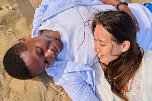 Young happy couple lying on the sand, top view. Loving multicultural couple, Asian woman and African man, faces close up. Love, relationship, beauty, romance, happiness, sensuality, people concept