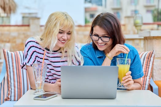 Smiling mother and teenage daughter looking in laptop screen together. Mom and teen girl resting together in outdoor cafe. Parent and child teenager, family, relationship, lifestyle, leisure concept