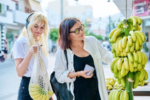 Mother and teenage daughter buying bananas at street retail market. With mesh reusable textile bag choosing farm organic natural fruits. Family, lifestyle, organic healthy food, tourism and travel