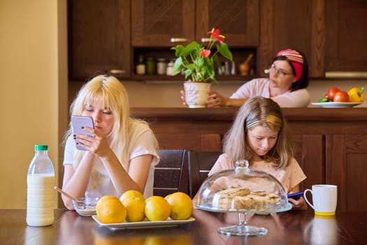 Family, girls children eating at table in kitchen. Home kitchen interior, mother preparing food, sisters eating and looking at smartphone screen. Lifestyle, daily routine, home, people, technology
