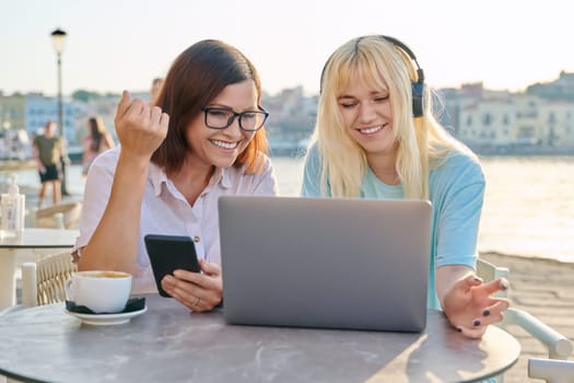 Happy smiling mom and teenage daughter looking at laptop together, sitting in outdoor cafe on seafront. Generation, vacation, parent teenager relationship, lifestyle, technology, communication, family