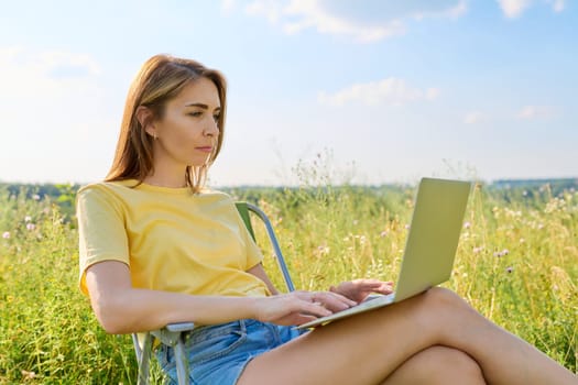 Adult woman sitting on folding camping chair using laptop, summer meadow with wild herbs background. Middle-aged female resting in nature, working remotely, freelance, lifestyle, technology concept