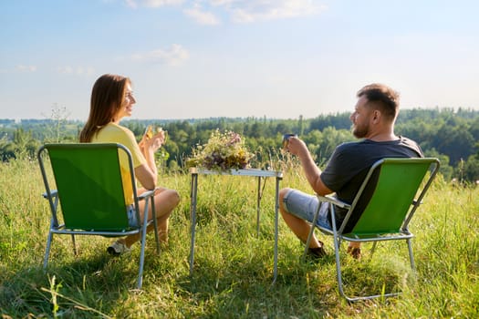 Happy middle-aged married couple having rest outdoors, in meadow, back view. Man and woman sitting on folding camping chair drinking coffee enjoying wildlife scenery sunbathing talking together