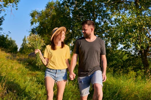 Happy romantic middle age couple, smiling man and woman walking together holding hands, female with bouquet of wildflowers, natural landscape in sunset. Relationships, love, dating, people 30s 40s age