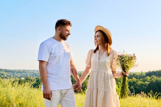 Couple holding hands smiling talking, summer nature blue sky background. Happy 40s age people, female with bouquet of flowers in dress hat, holiday, vacation, outdoor walk, relationship concept