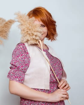 A red-haired woman in a fashionable outfit playfully looks out from behind a reed