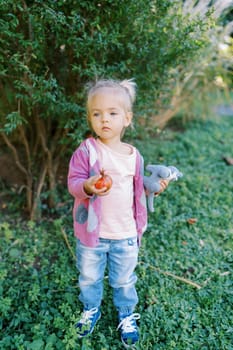 Little girl with a ripe pomegranate and a toy in her hands stands near a green bush in the garden. High quality photo
