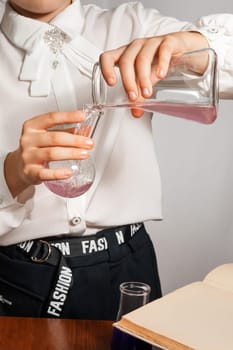 A schoolgirl conducts a chemical experiment based on a textbook. Pours pink liquid from one flask into another close-up