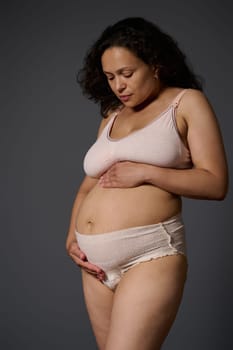 Studio portrait of new mom, a multi ethnic middle aged woman, touching her belly, isolated on gray background, dressed in lingerie and showing her body with stretch marks and postpartum flaws