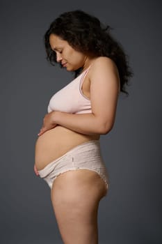 Studio side portrait of a woman new mother in underwear, showing her postpartum belly with stretch marks and flaws, isolated on gray background. Post pregnancy and maternity lifestyle. Body positivity