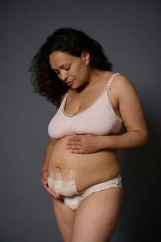 Confident authentic portrait of a multi ethnic middle aged woman, new mother touching her belly with a bandage hiding scars after c-section, few days after childbirth, isolated on gray background