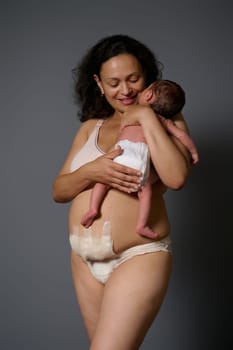 Happy mother in lingerie, showing naked belly with bandage hiding scars after c-section cesarean and postpartum stretch marks, smiling while holding her newborn baby, isolated gray studio background