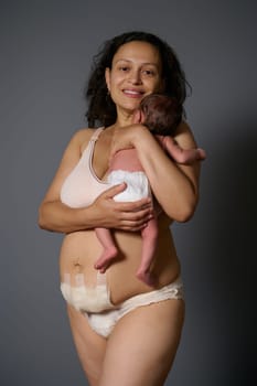 Happy new mother smiles looking at camera, holds her newborn baby and shows naked belly with bandage hiding scars after c-section and postpartum flaws like stretch marks, isolated over gray background
