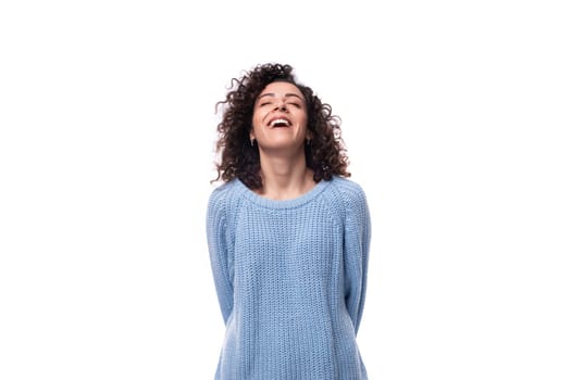 authentic smiling young brunette woman with curls is dressed in a blue knitted sweater on a white background.