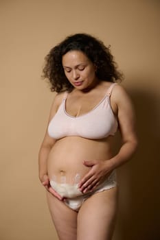 Confident authentic woman mother holding her postnatal belly, with bandage hiding scar after C-section cesarean, showing stretch marks and her tummy after childbirth, isolated beige studio background
