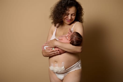 Authentic studio portrait charming woman in underwear, smiles looking at her newborn baby she's holding, standing on beige background, showing her body with postpartum flaws and scar after C-section