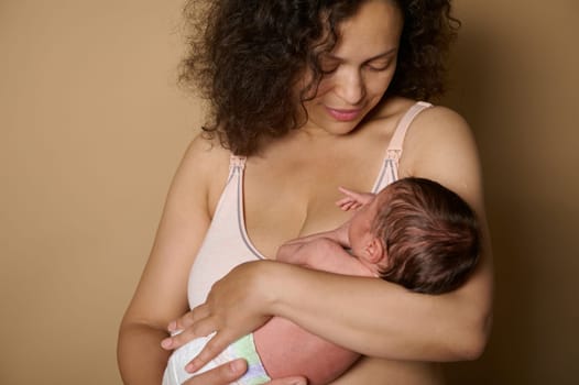 Close-up portrait of a happy multi ethnic pretty woman, loving caring happy mother smiling looking at her newborn baby, she hold in her arms, isolated on beige studio background. Postpartum. Maternity