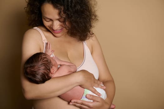 Happy young mother expressing positive emotions holding her new born baby, standing naked over beige studio background, showing her postnatal body with flaws few days after the childbirth. Postpartum