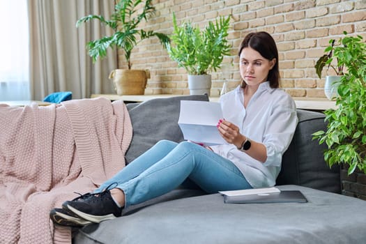 Serious young woman reading official paper letter while sitting on sofa in living room at home. Notification from bank, work, social tax services, invoice, insurance company, about rental property