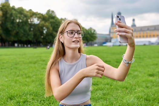 Teenage girl student having video call on smartphone, resting sitting on grass lawn near university building. Technology, leisure, communication, youth