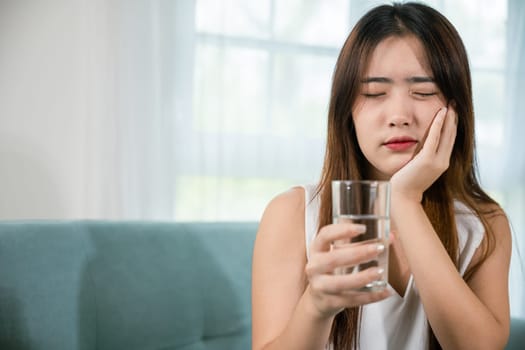 Asian young woman with sensitive teeth holding glass of cold water at home in living room, beautiful female toothache and dental problems touching cheek feeling pain after drinking cold water