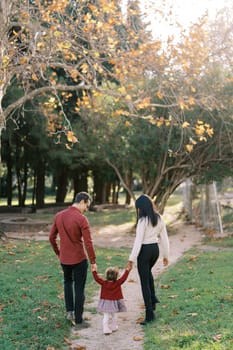 Mom and dad with a little girl walk along the path in the park, holding hands. Back view. High quality photo