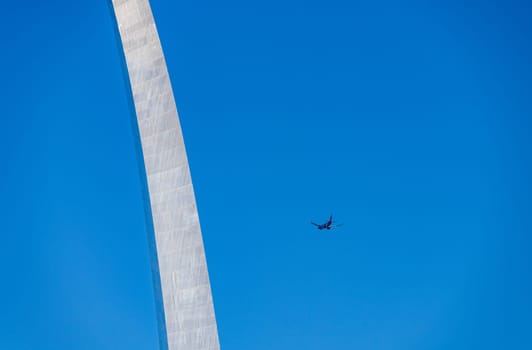 Simple abstract view of the Gateway Arch in St Louis set against a plain blue sky with landing aircraft approaching the airport