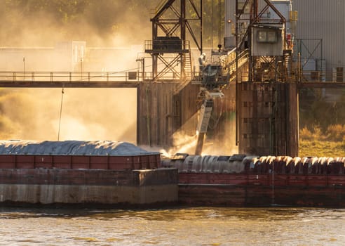 Tugboat pushing freight barges past grain loading dock with dust illuminated by the sun in East St Louis Illinois