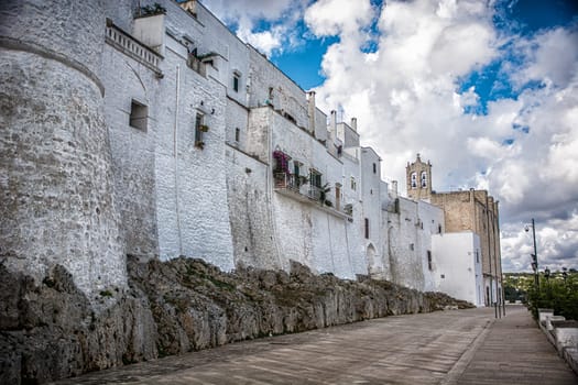 A view of Ostuni, Italy, the White City nestled on a hill overlooking the Adriatic Sea in Puglia, Italy. The photo shows a row of white buildings sitting on top of a hill, with the Adriatic Sea in the distance. The buildings are all different shapes and sizes, but they are all painted white, which gives the town its unique appearance.