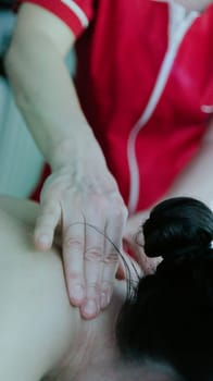 One young Caucasian unrecognizable masseur in a red robe gives a girl a relaxing massage on her neck using her fingers in a home beauty salon, close-up side view.