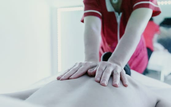 One young Caucasian unrecognizable masseur in a red robe gives a girl a relaxing massage on her back using her palms in a home beauty salon, bottom side view close-up.