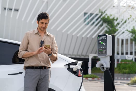 Young man use smartphone to pay for electricity at public EV car charging station at city commercial mall parking lot. Modern environmental and sustainable urban lifestyle with EV vehicle. Expedient
