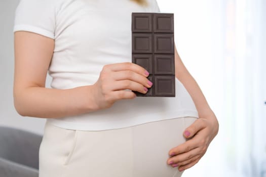A pregnant woman is holding a bar of chocolate and cradling her stomach. Sweets during pregnancy concept