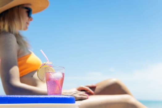 Happy young european girl drinks a cocktail and rests in a sun lounger on the beach, space for an inscription portable blue refrigerator by the sea. High quality photo