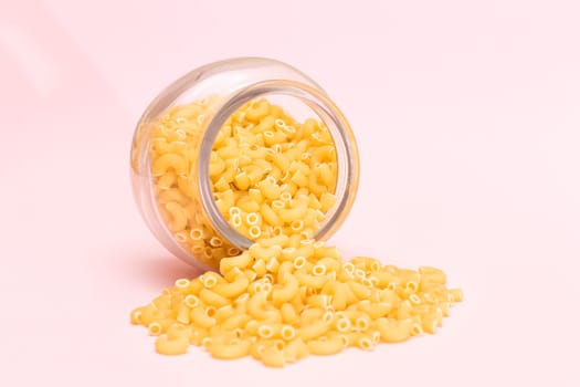 Uncooked Chifferi Rigati Pasta in Glass Jar on Pink Background. Fat and Unhealthy Food. Scattered Classic Dry Macaroni. Italian Culture and Cuisine. Raw Pasta