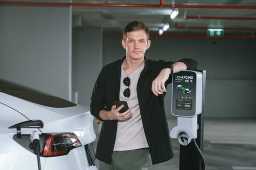 Young man travel with EV electric car to shopping center parking lot charging in downtown city showing urban sustainability lifestyle by green clean rechargeable energy of electric vehicle innards