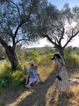 Girl-photographer shoots a kissing man and woman sitting on the grass near a tree. High quality photo