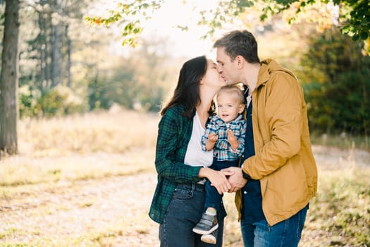 Mom and dad are kissing while holding their little daughter in their arms in the autumn park. High quality photo