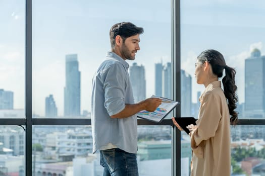 Professional businessman present financial statistic to investor while standing near panoramic window shows skyscraper or city view. Diverse businesspeople discuss about marketing plan. Tracery.