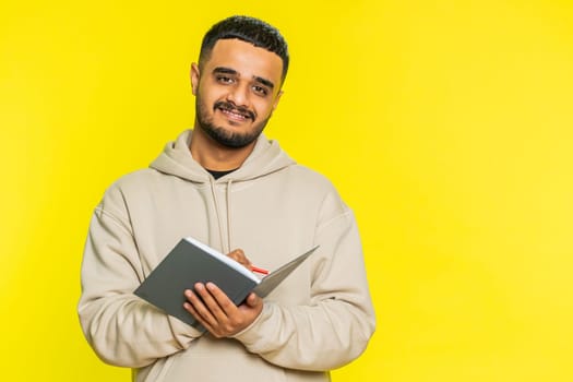 Thoughtful journalist young Indian man making notes, writing down thoughts with pen into notepad notebook diary, to do list, good idea inspiration. Arabian Hindu guy isolated on yellow background